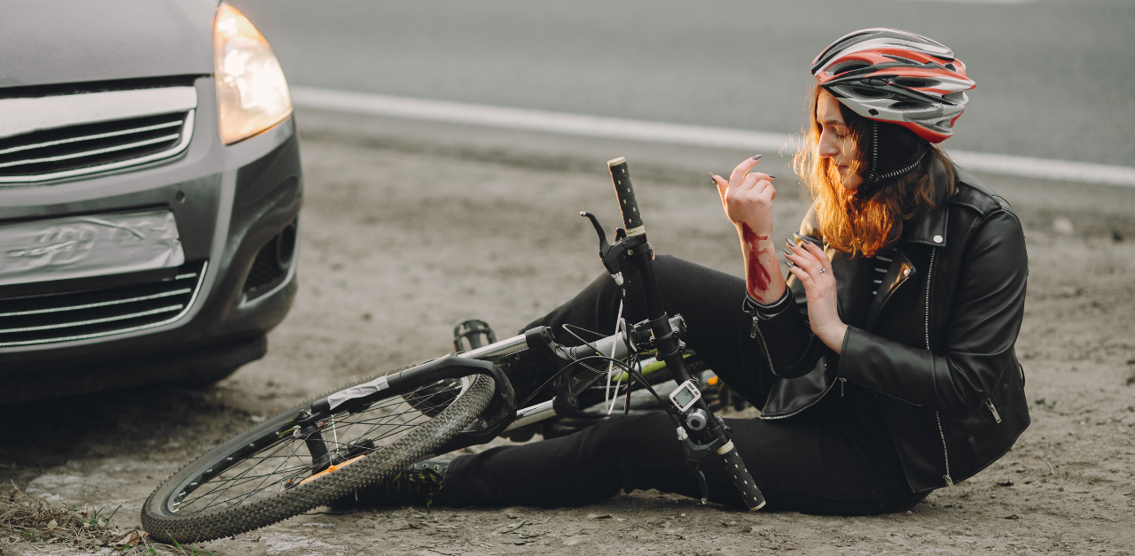 Woman wearing a helmet and black jacket sits on the roadside next to a bicycle, examining a scrape on her arm, with a car nearby