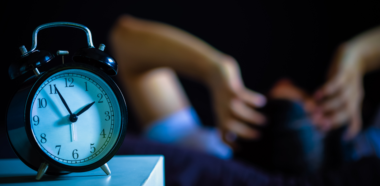 A close-up of an alarm clock showing 5:50 AM, with a blurred figure holding their head in the background, suggesting stress or insomnia