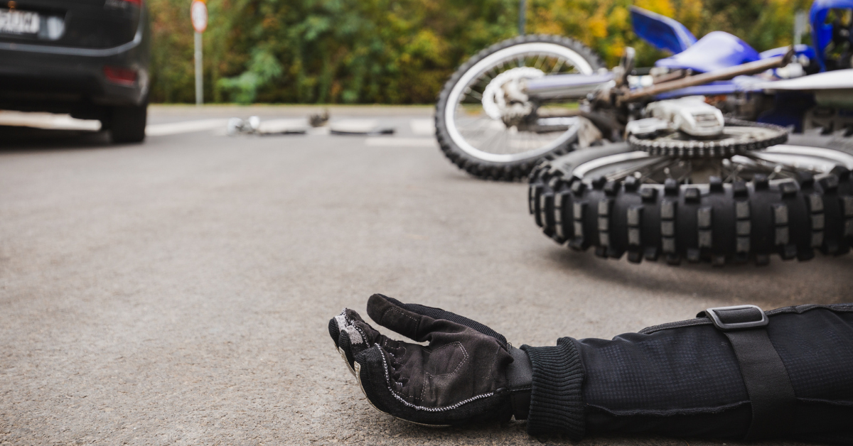 A motorcyclist's gloved hand lies on the road near a fallen motorcycle, with a car in the background, suggesting a crash scene