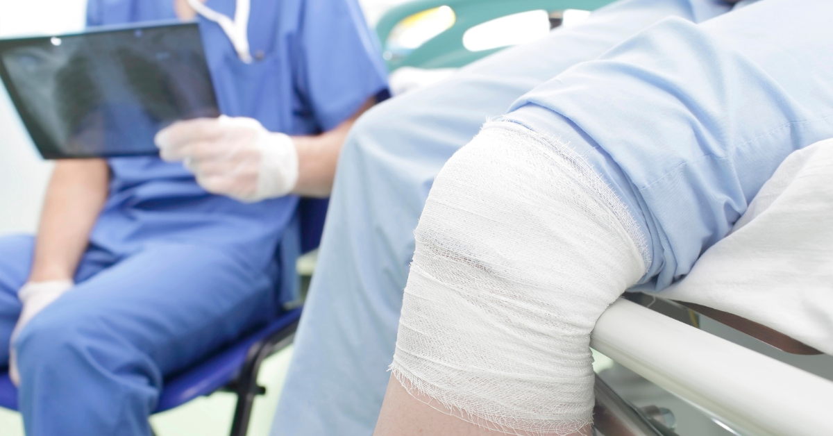 A close-up of a patient's bandaged knee in a hospital bed, with a nurse in scrubs reviewing an X-ray in the background