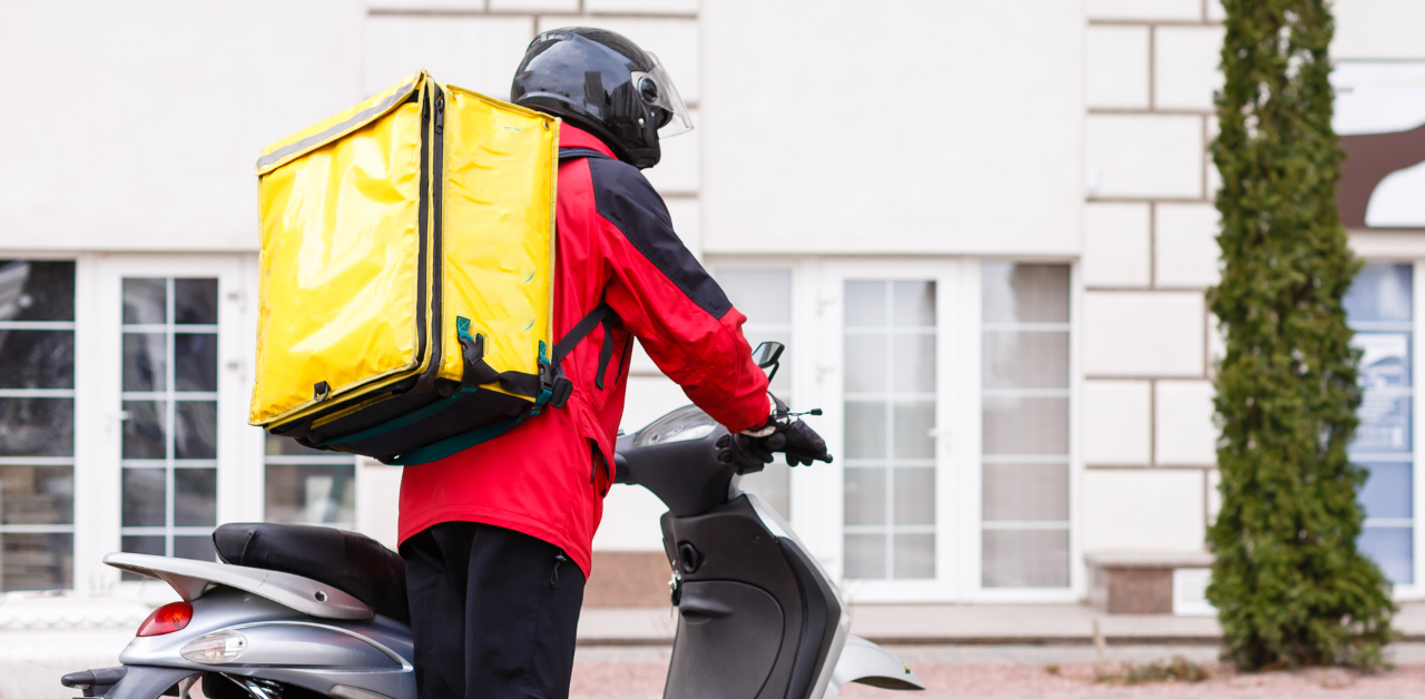 Food delivery driver on a scooter wearing a helmet and red jacket with a large yellow insulated backpack