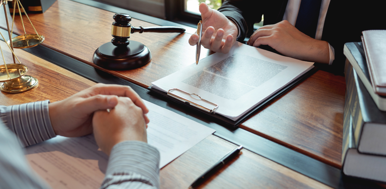 Lawyer discusses case details with client at desk, legal documents, gavel, and scales in view