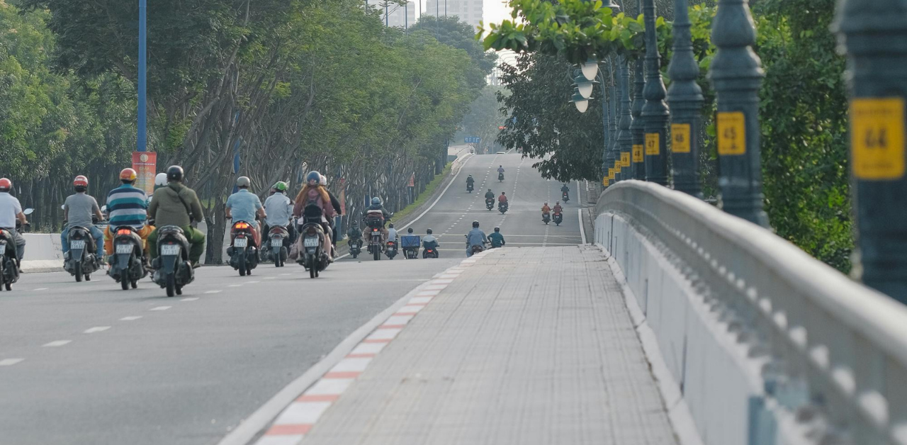 Dozens of motorbikes travel on a wide, tree-lined urban road under clear skies and light traffic flow