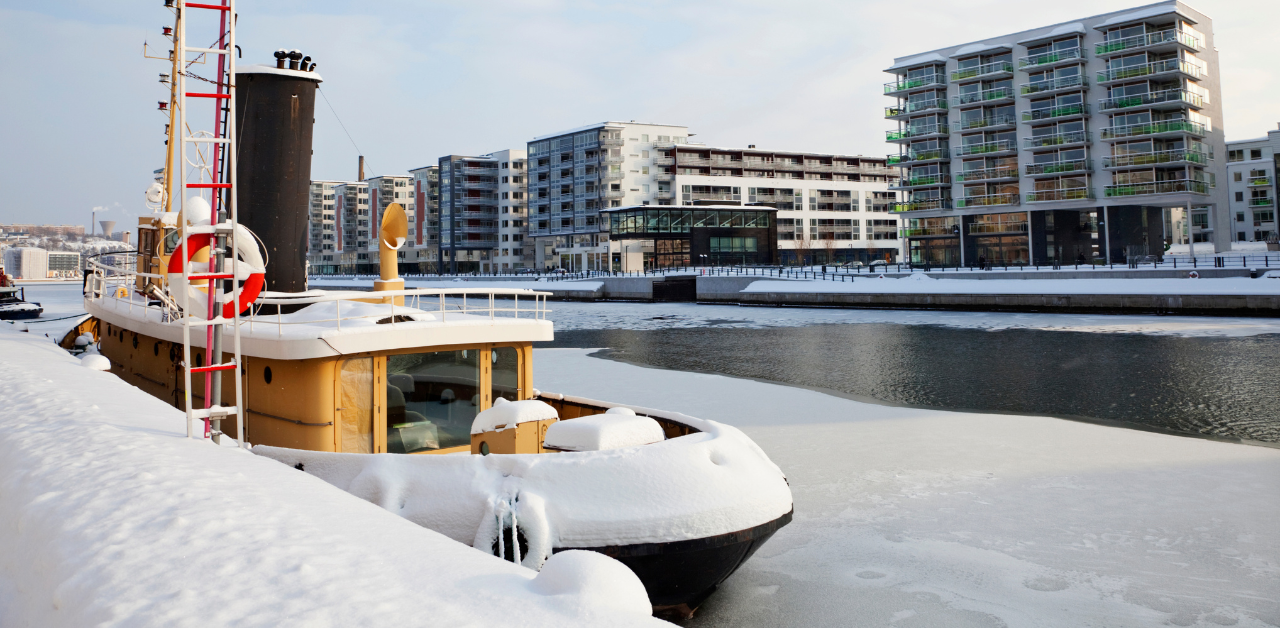 Snow-covered boat docked in a frozen urban harbor surrounded by modern apartment buildings