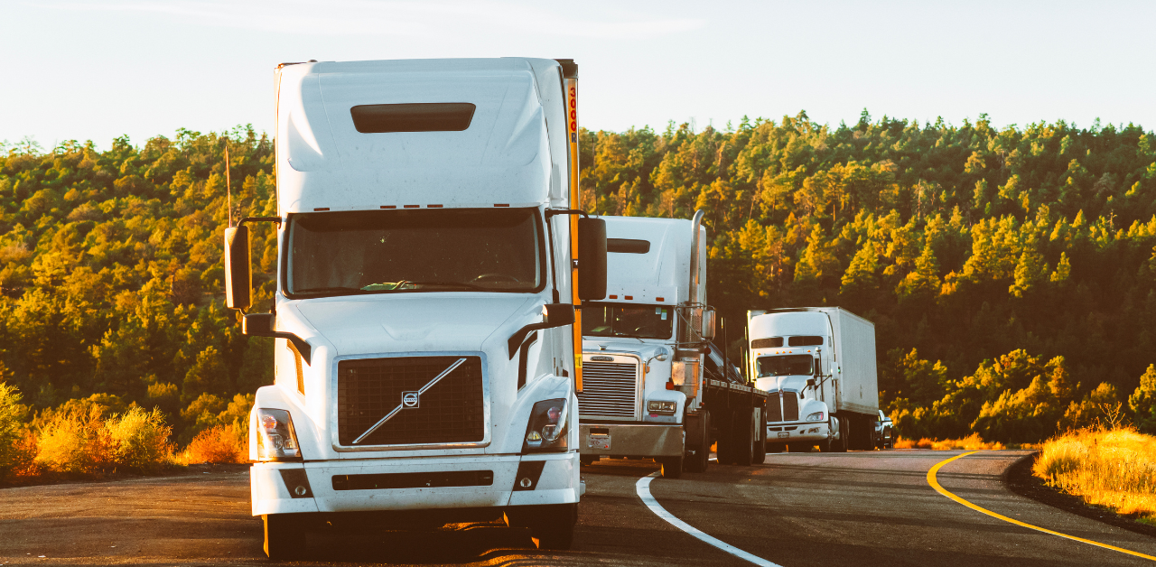 Three white semi-trucks driving on a curved highway surrounded by green forest