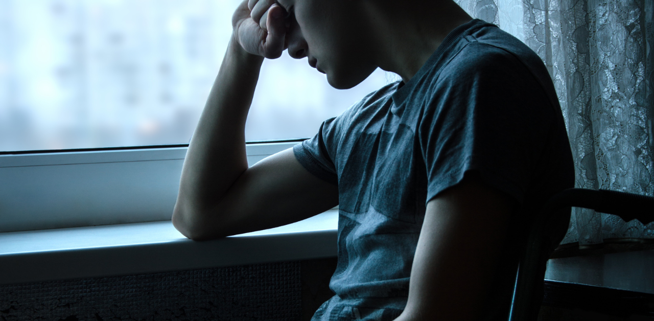 Teen boy sits alone by window, head in hand, expressing signs of depression or anxiety