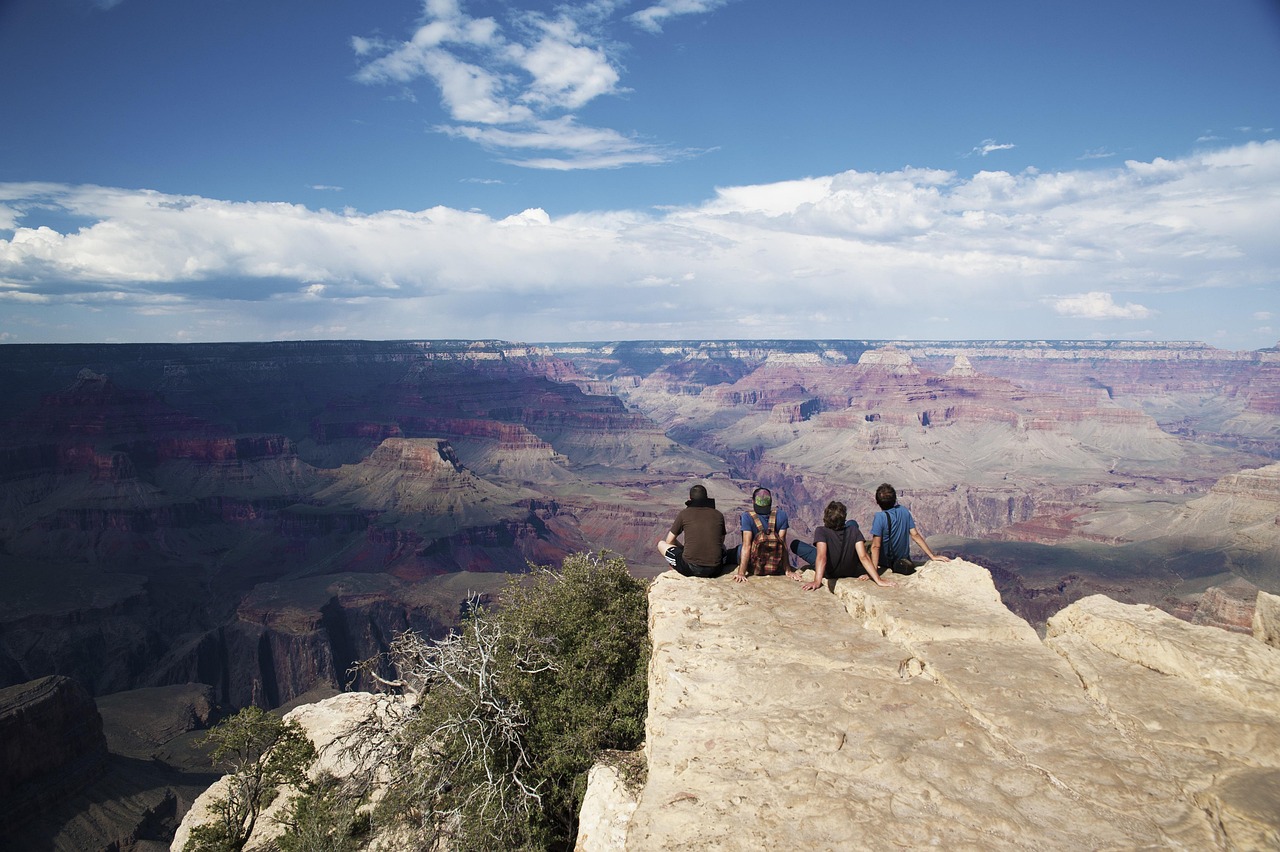 Group of friends sitting on a cliff edge, admiring the vast Grand Canyon under a clear blue sky