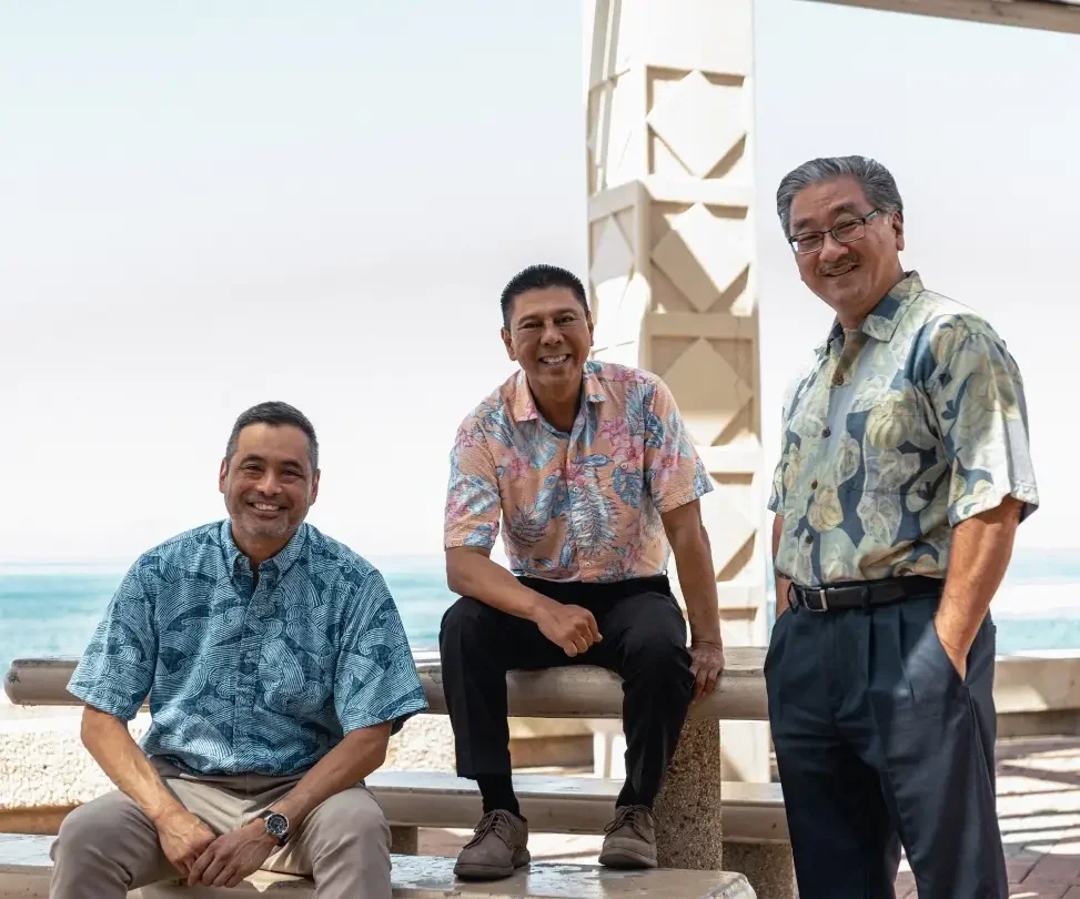 Three Hawaiian men in patterned shirts smiling, with a beach and white structure in the background.