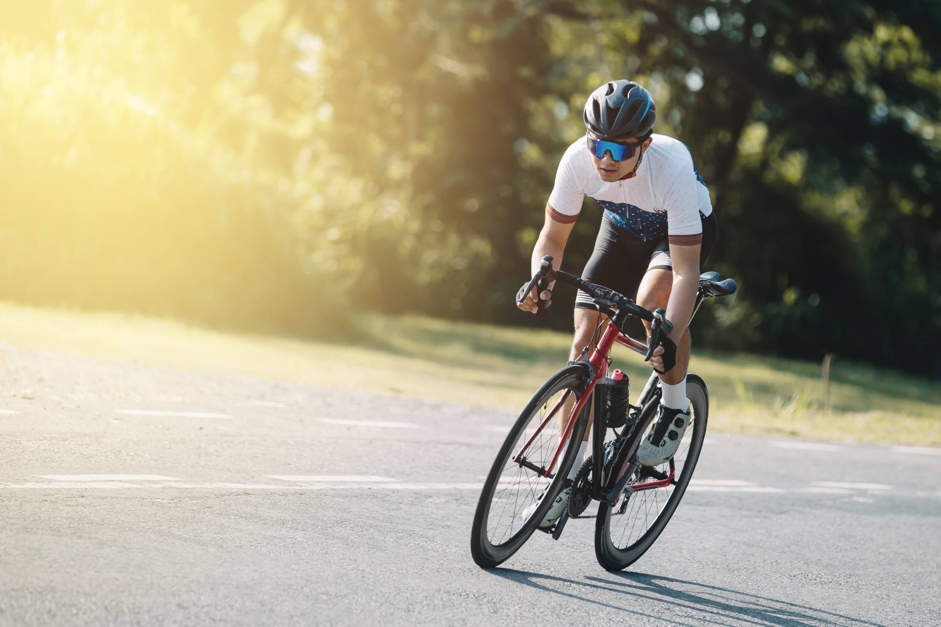 A male cyclist in a helmet and sunglasses rides a red and black road bike on an empty paved road with trees in the background.