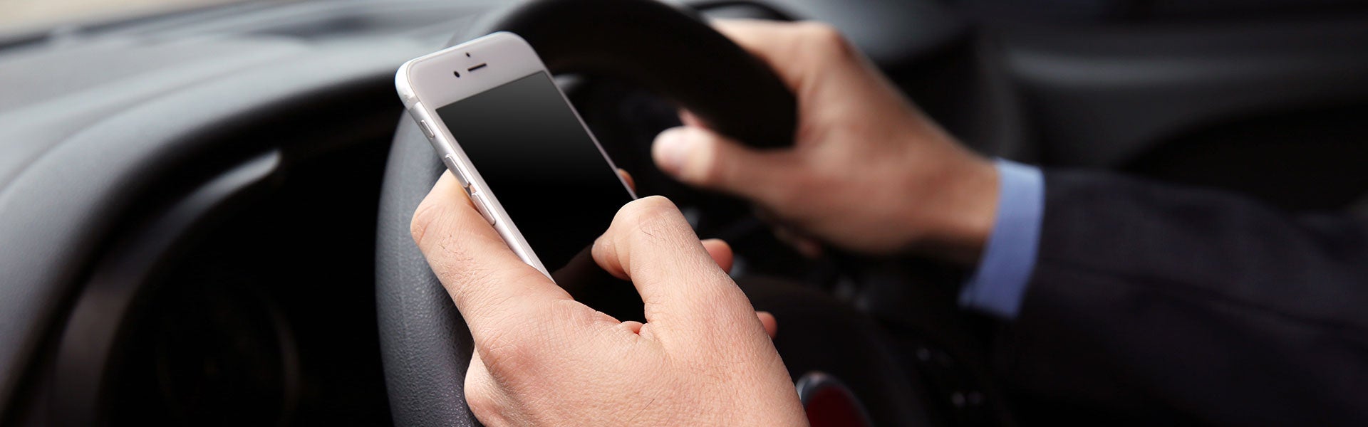 Close-up of a person holding a smartphone with their left hand and steering wheel with their right hand while driving.