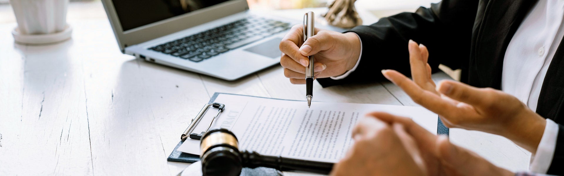 A person in a black suit holds a pen over a contract, with a laptop and gavel nearby.
