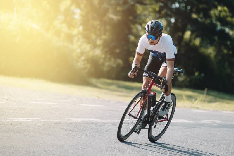 A person in cycling gear, glasses, and a helmet rides a racing bike on a sunlit road, focused on the ride.