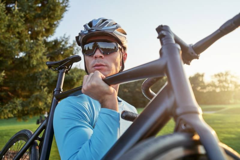 Cyclist in helmet and sunglasses carries a black road bike over their shoulder outdoors on a sunny day.