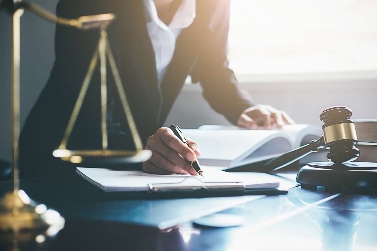A lawyer writes, a gold scale of justice and a gavel on the table, reading a book in a sunlit office.