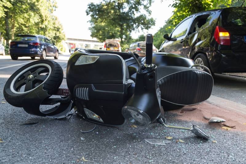 A black scooter lies damaged on the road after a crash, next to a black car with a broken window.