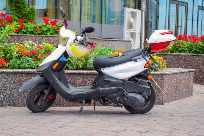 A white and black scooter with a yellow helmet hanging on the handlebars, parked on a brick sidewalk.