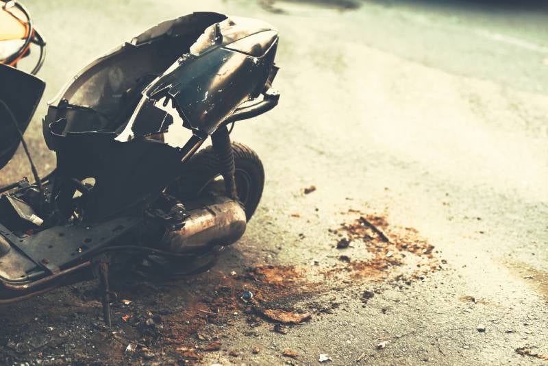 A heavily damaged, black scooter laying on its side on gray asphalt with rust-colored debris scattered around it.