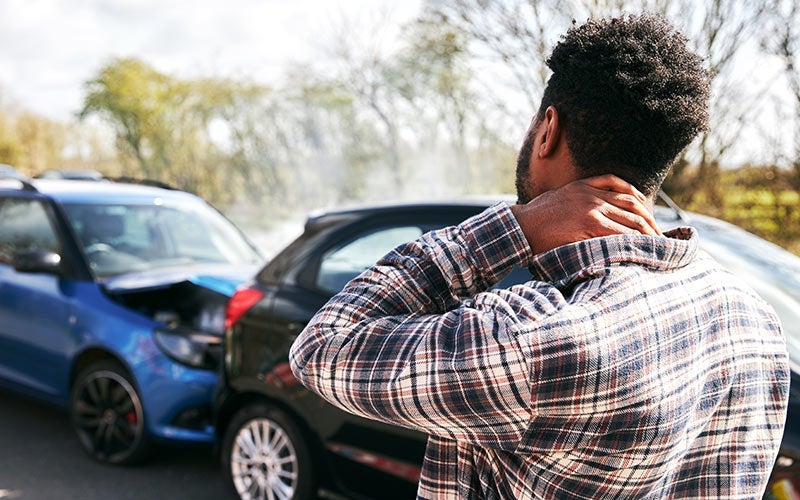 A man from behind, hand on his neck, looking at two cars crashed on the road, with smoke rising from one car.