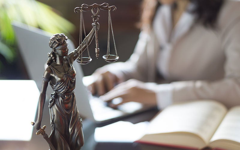 Lady Justice statue with scales and sword. Blurry background shows person typing on laptop and open book.
