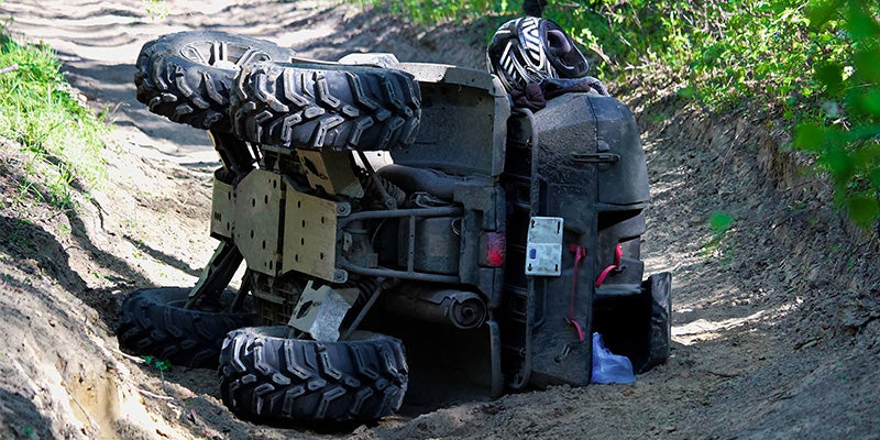 An ATV is overturned on its side in a dirt trail, with a helmet resting on its highest point.