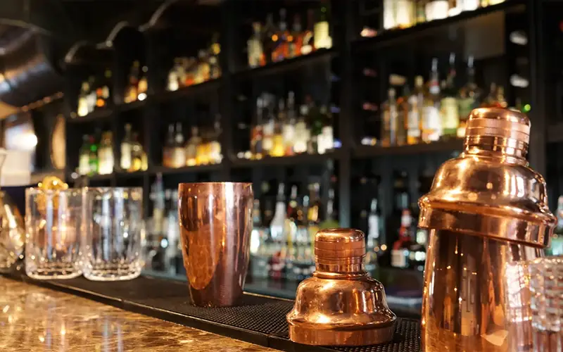 Close-up of copper bar tools and empty glasses on a marble bar, with blurred liquor bottles in the background.