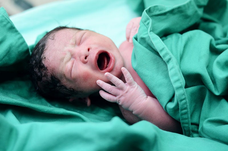 Newborn baby with eyes closed, mouth open wide, and tiny hand visible, wrapped in a green blanket.