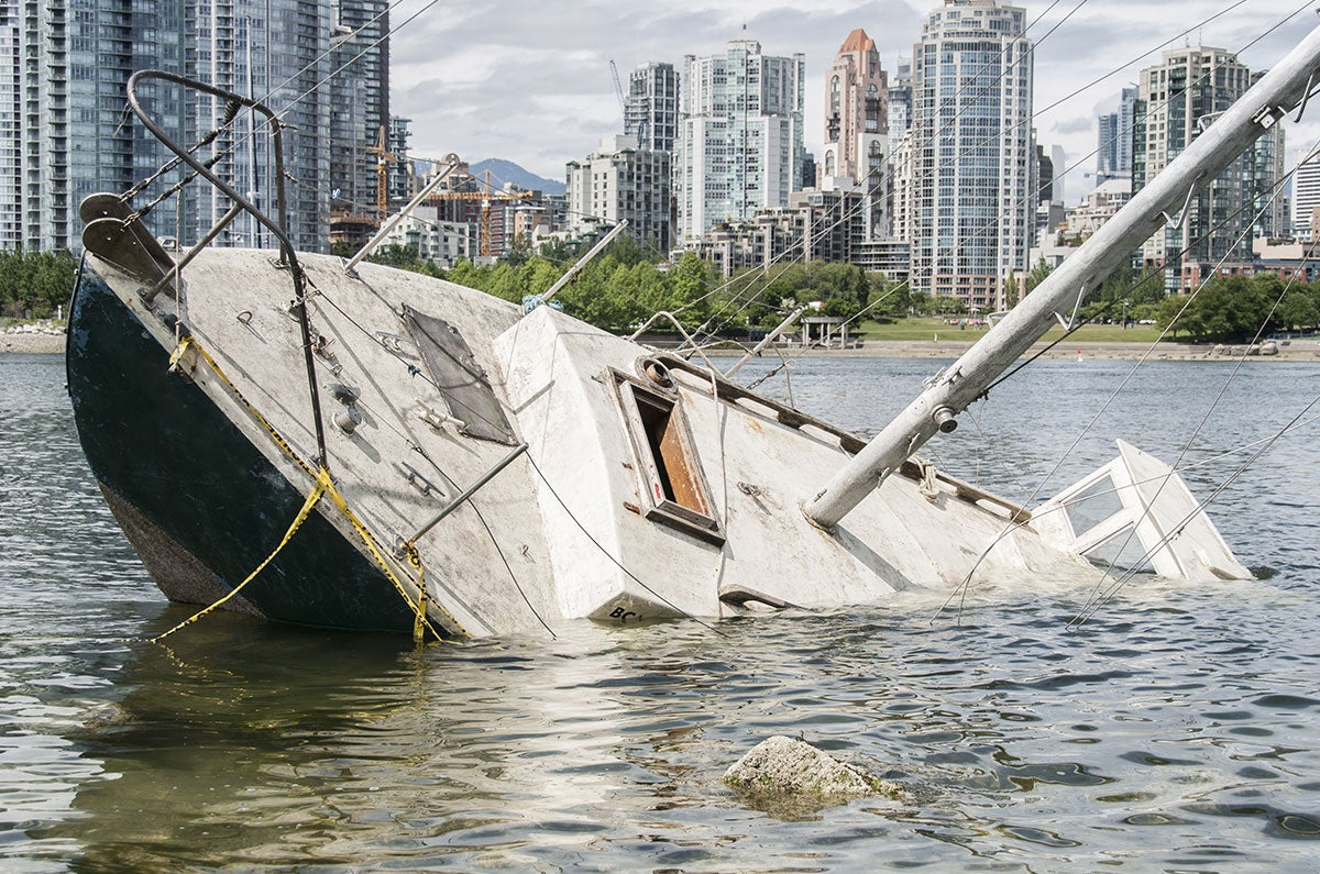A partially sunken white sailboat with green trim in the foreground of a city skyline with tall buildings.