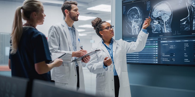 Three medical professionals examining brain scans on a large display, with one pointing to an image.