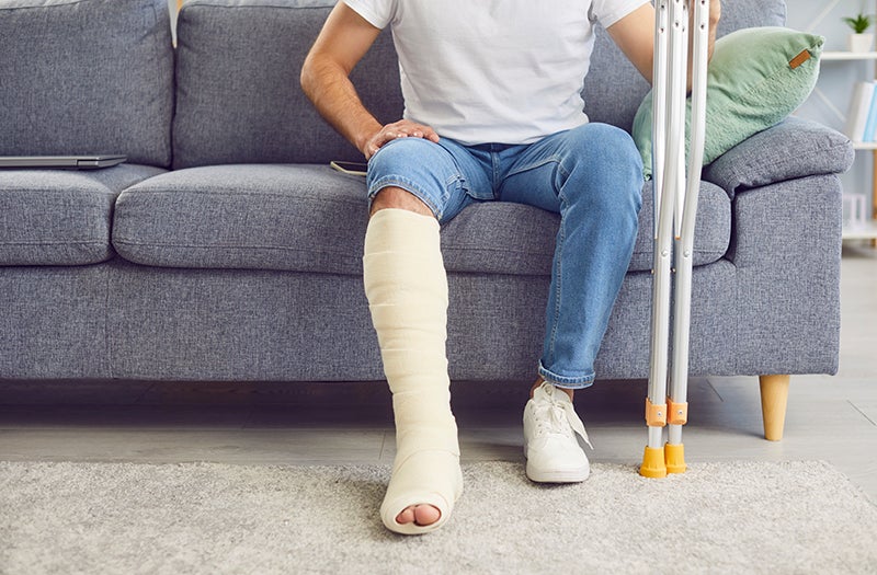 A man with a white t-shirt and blue jeans sitting on a gray sofa, holding crutches, with a bandaged leg.
