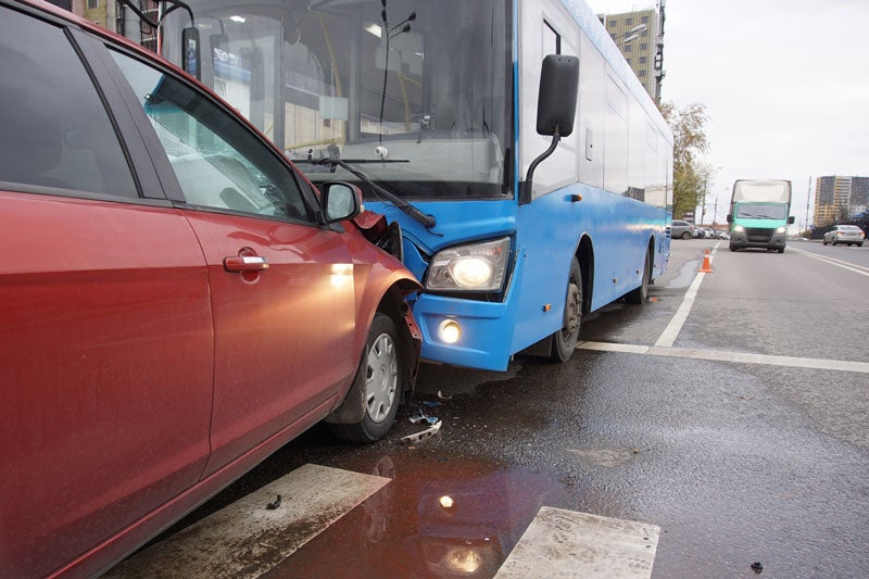 A red car crashed into the front, passenger side of a blue bus at a crosswalk on a wet road.