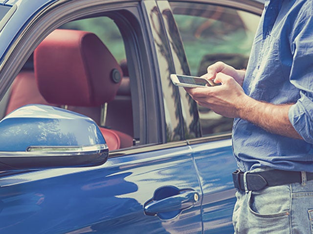A man in a blue shirt uses his smartphone next to a parked blue car with a red interior.