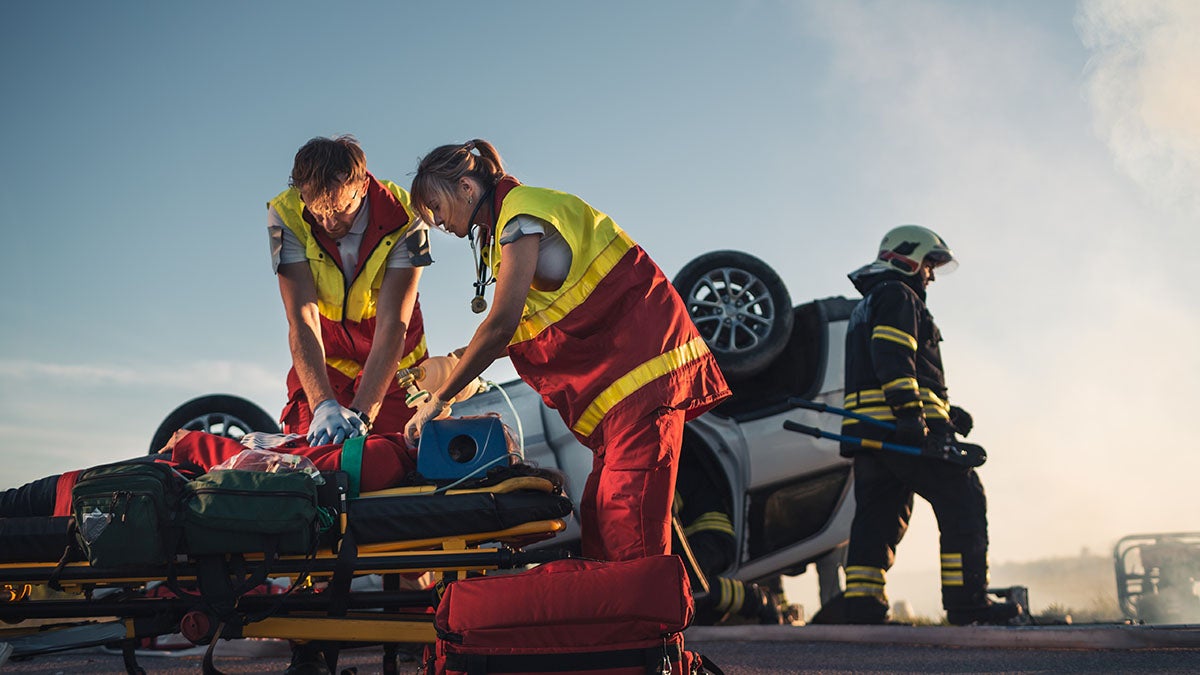 Paramedics and a firefighter attend to an injured patient on a stretcher beside an overturned car.