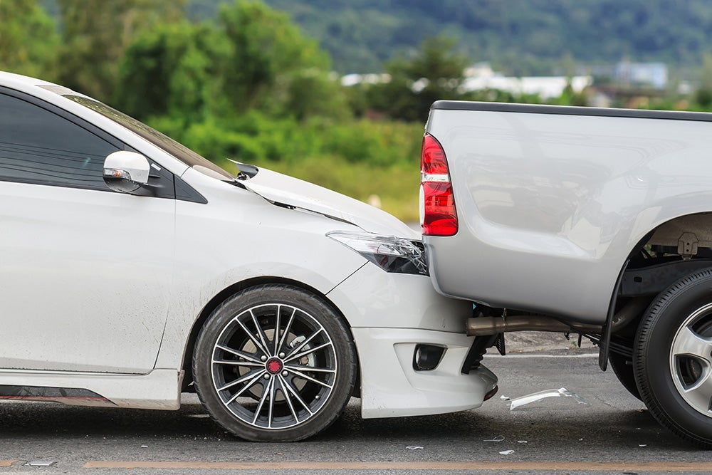 A white sedan rear-ends a silver pickup truck, causing visible damage to the sedan's hood and bumper.
