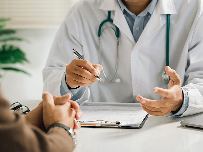 A doctor in a white coat and stethoscope talking to a patient, holding a pen over a medical document.