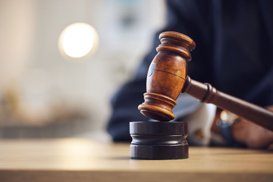 A wooden gavel resting on its block, with a person in a dark suit partially visible in the blurred background.