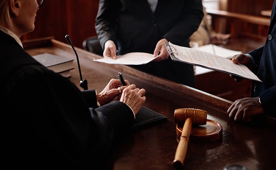Judge signing papers on a courtroom bench with a gavel nearby, two other people holding documents.