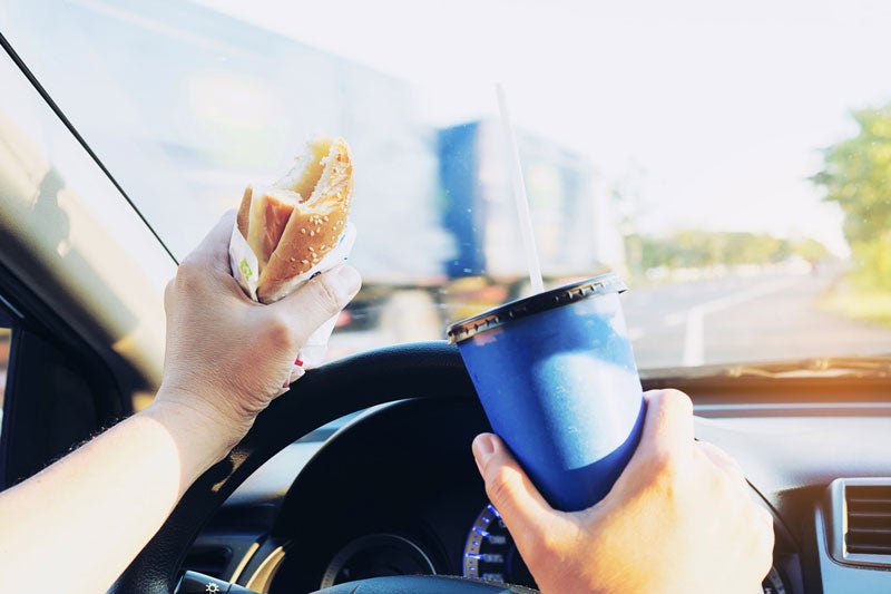 Hands holding a half-eaten burger and a blue soda cup while driving, with a highway visible through the windshield.