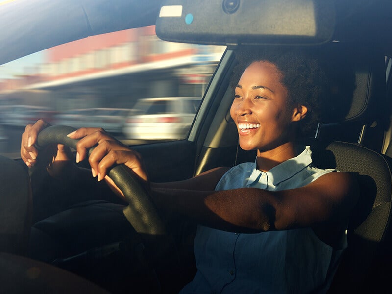 A smiling woman with sun on her face drives a car, hands on the steering wheel, with blurred scenery outside.