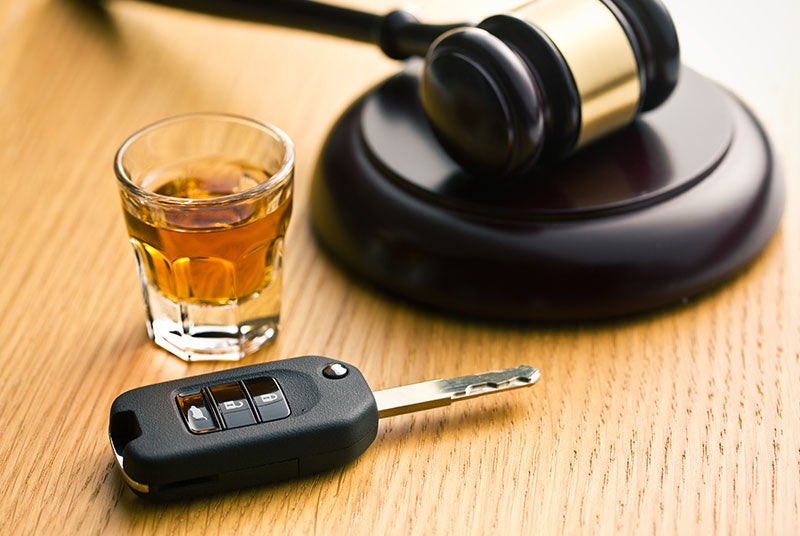 A shot glass of amber liquor, a car key, and a judge's gavel on a wooden surface.