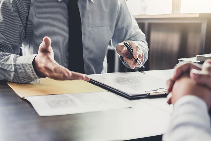 A professional in a gray shirt and dark tie gestures with one hand at a document on a clipboard and holds a pen in the other, across a desk from another person.
