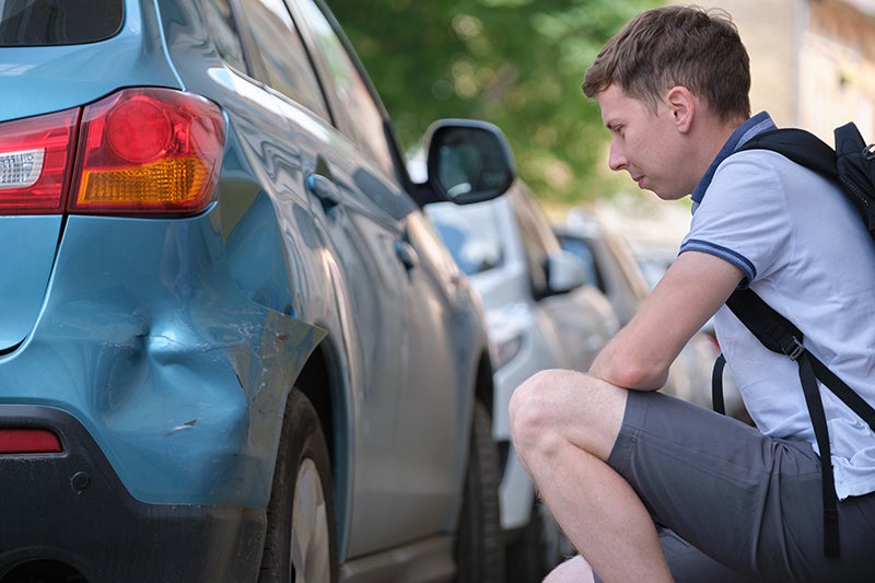A worried man looks at the dented and scratched rear bumper of a light blue car.