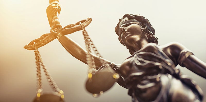 A bronze statue of Lady Justice, blindfolded and holding scales, with a bright sun flare.