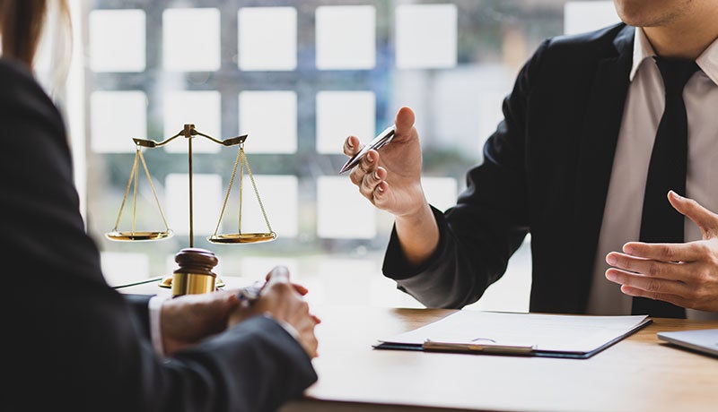 Two lawyers, one speaking and gesturing with a pen, sit across a table from each other in front of scales of justice.