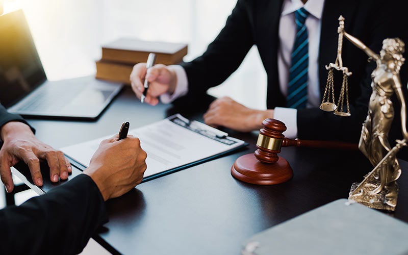 Two legal professionals at a desk, one signing a document, with a gavel and a Lady Justice statue nearby.