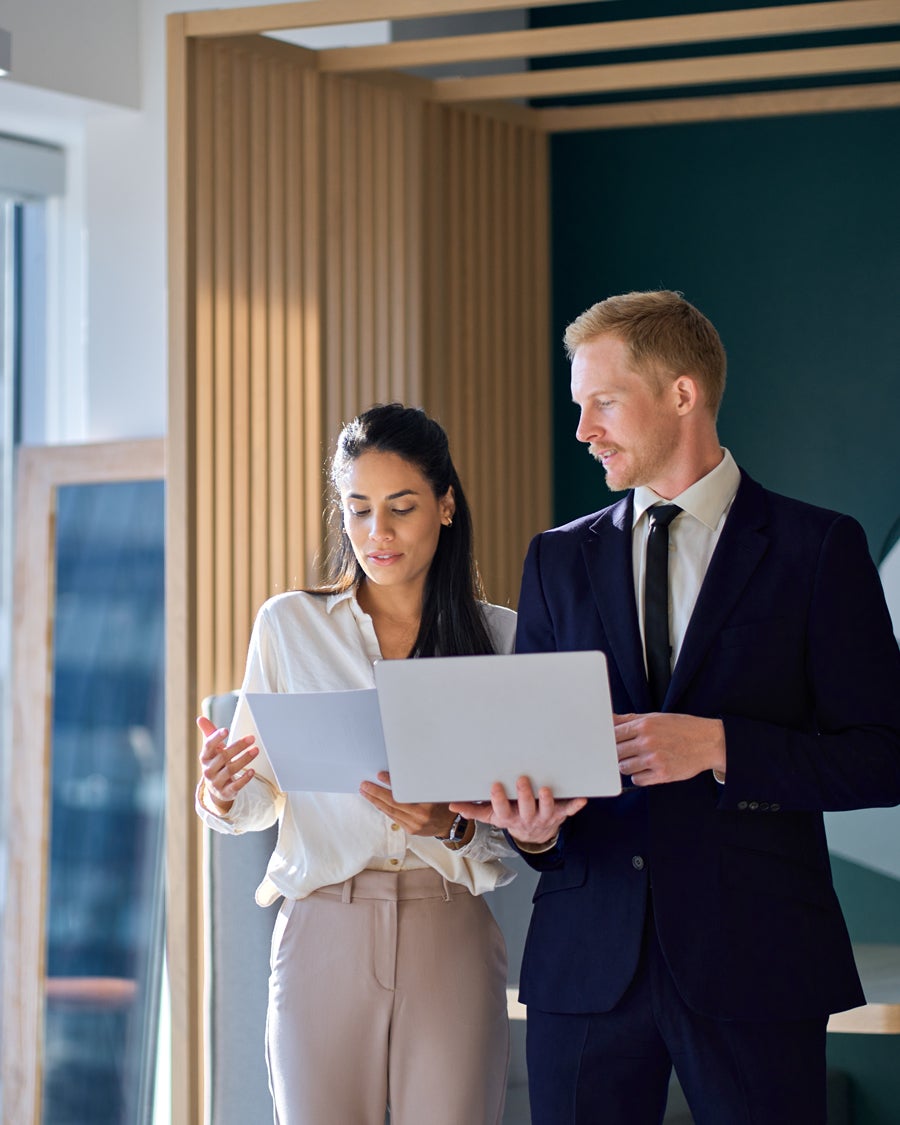 Two professionals, a woman with documents and a man with a laptop, discuss work in a modern office space.