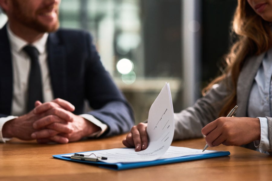 A woman signs a document on a blue clipboard with a man in a suit opposite her at a wooden table.