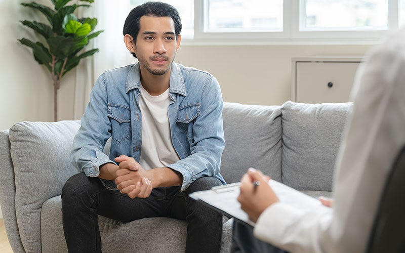 A young man sits on a gray couch, looking thoughtfully at a person taking notes on a clipboard.