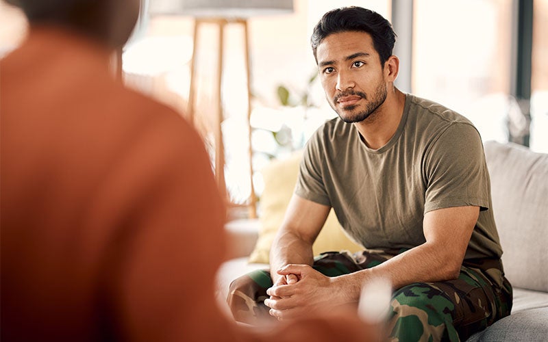 A man in a military uniform sits on a couch, listening intently to someone across from him.