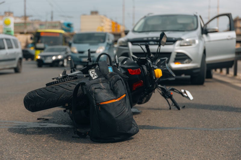 A black motorcycle and backpack lie on the pavement after a traffic accident with a silver SUV and other cars.