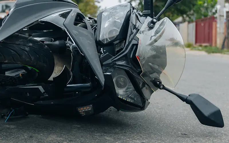 A crashed dark grey motorcycle lying on its side on a paved road, front headlight and tire visible.
